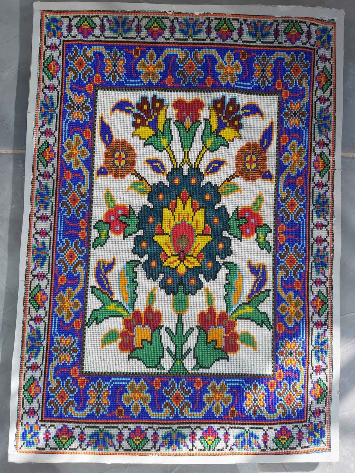 Unframed handmade bead artwork: a yellow lotus-like flower in a dark teal medallion with a bouquet of crimson flowers on a white ground, inside cobalt-blue and white floral borders.
