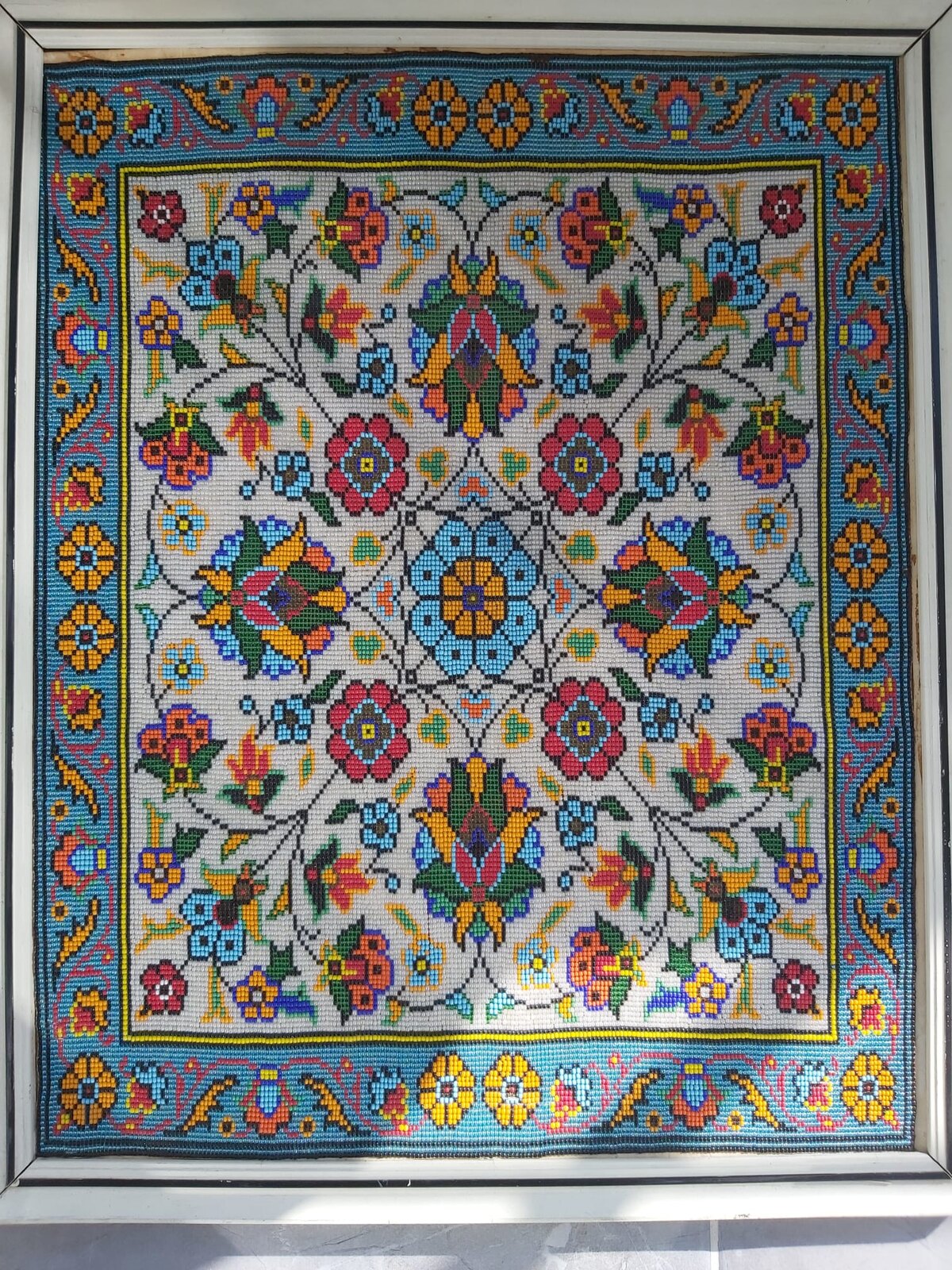 Framed handmade bead artwork with a symmetrical floral design: a sky-blue central rosette, crimson flowers and orange palmettes linked by black vines on an ivory ground, inside a wide turquoise border of orange and yellow rosettes.